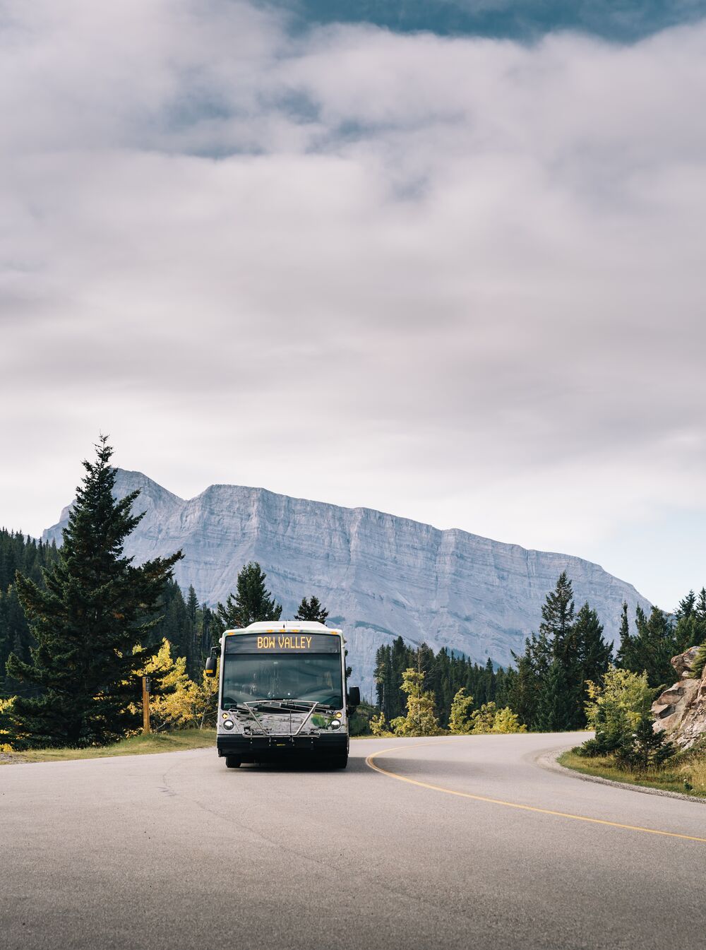 A Roam Regional Transit bus drives on the Minnewanka Loop in Banff National park with Mount Rundle in the distance behind it.